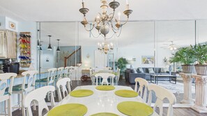 Amplia sala de estar con una araña, comedor con sillas blancas y individuales verdes, y cocina con botellero.