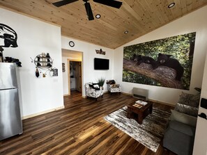 A spacious living room with a wooden ceiling, a large bear-themed wall mural, a modern kitchen area, and a cozy seating arrangement. - Country Quite 1 Bedroom, Looks like a cabin on the outside but modern inside (Johnson)