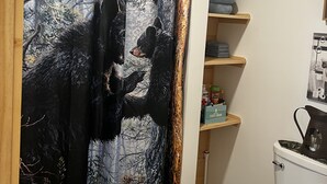 A bathroom with a shower curtain depicting bears, wooden shelves, a toilet, and a trash bin.