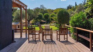 A wooden deck with chairs and a table, overlooking a garden with various plants and a building in the background.