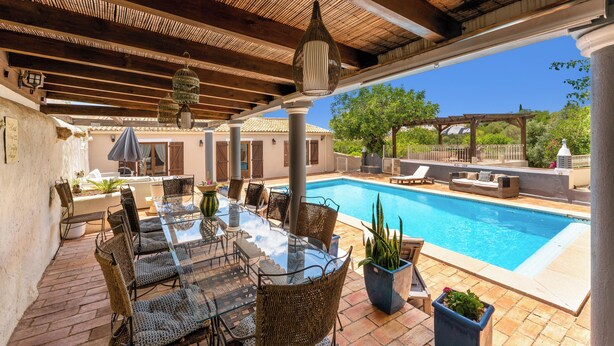 An outdoor pool area with a glass table and wicker chairs, surrounded by potted plants and a pergola.