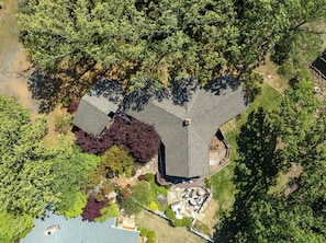 An aerial view of a house with a large, sloped roof, surrounded by dense trees and a well-maintained lawn.