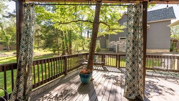 Une terrasse extérieure couverte, dotée d’une rambarde en bois, d’une plante en pot et offrant une vue sur une maison en arrière-plan.