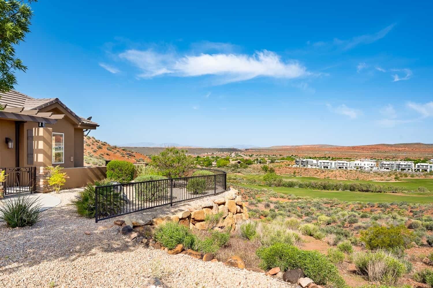 A modern house with a stone pathway leading to a gated entrance, surrounded by desert vegetation and a view of a residential area in the distance.