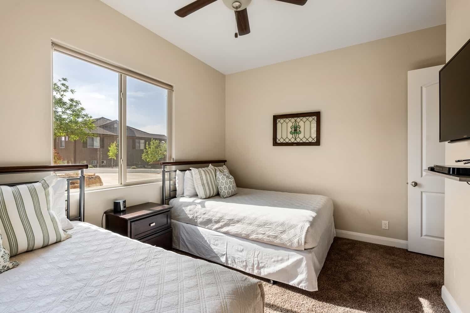A bedroom with two beds, a ceiling fan, a window with a view of buildings, and a door with a mounted TV.