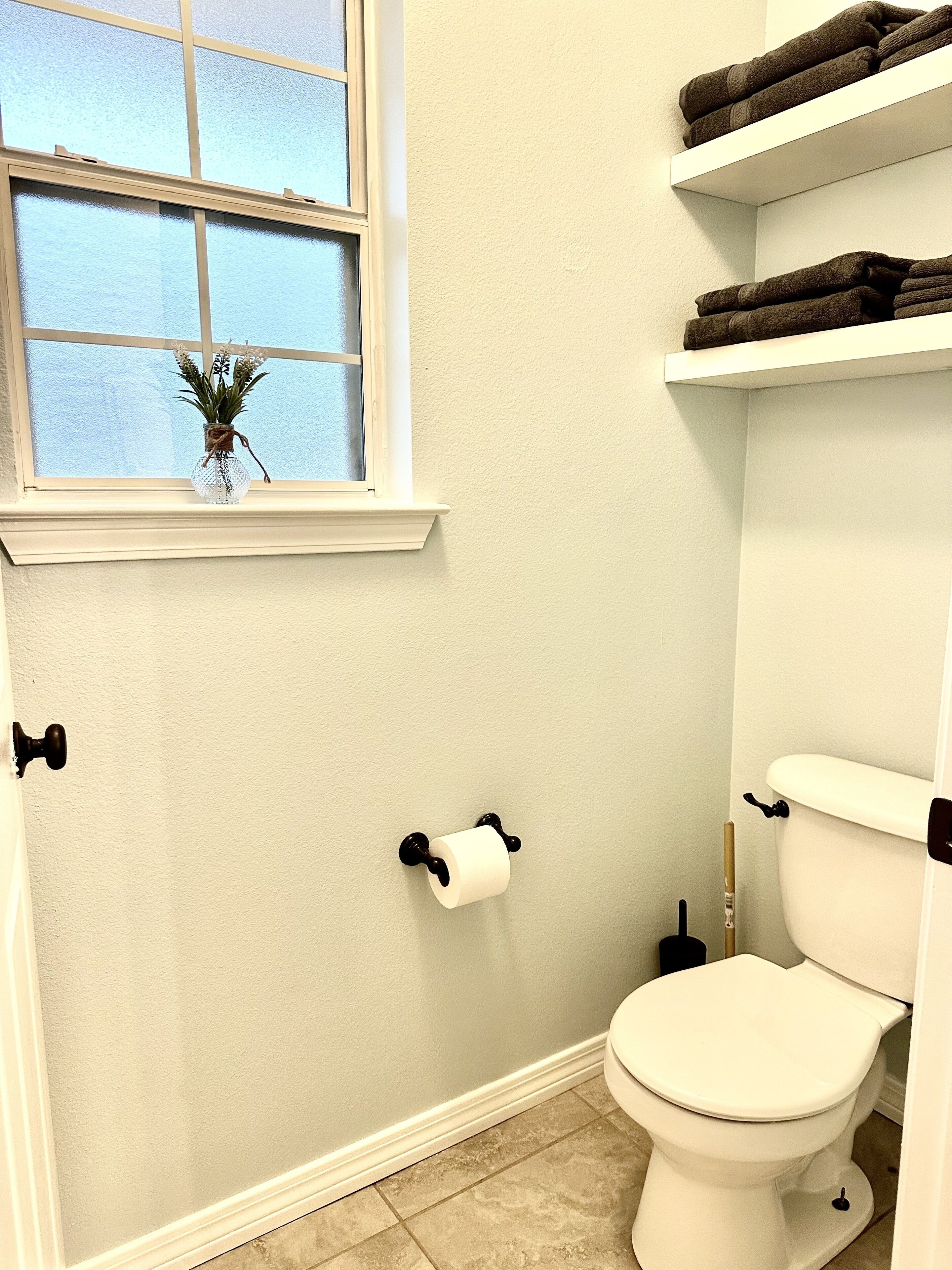 Un baño con un inodoro, un rollo de papel higiénico, una ventana con una planta y repisas con toallas.