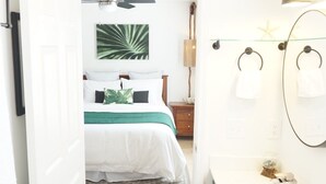 A bedroom with a bed, a bedside table, and a nightstand. There is a bathroom with a sink, mirror, and towel rack. - Emerald Hideaway 2 Bedroom Condo by RedAwning (Navarre)
