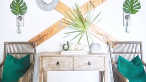 A room with a wooden console table, two wicker chairs, a large plant, and wall decorations including a wooden cross, a mirror, and two tropical leaves. - Emerald Hideaway 2 Bedroom Condo by RedAwning (Navarre)