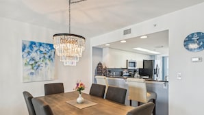 A dining area with a wooden table, six chairs, a chandelier, a painting, and a kitchen in the background.