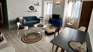 A modern living room with a blue sofa, a dining table with chairs, a coffee table, and a TV. - Appartement Neuf 83m² vue Mer, Pleins Coeur de St Florent (Saint-Florent)