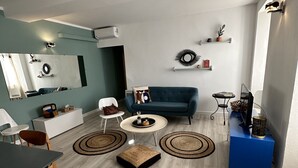 A modern living room with a blue sofa, white chairs, a coffee table, and a dining area with a table and chairs. - Appartement Neuf 83m² vue Mer, Pleins Coeur de St Florent (Saint-Florent)