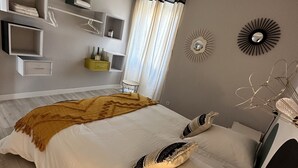 A bedroom with a large bed, built-in shelves, a bedside table, and a window with curtains. - Appartement Neuf 83m² vue Mer, Pleins Coeur de St Florent (Saint-Florent)