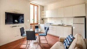 Una cucina moderna con mobili bianchi, forno a incasso e frigorifero. C'è una zona pranzo con un tavolo e sedie in vetro, e una TV montata a parete.