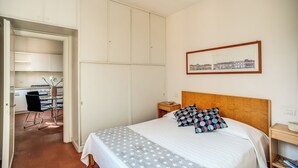 Una camera da letto con un letto, una testiera in legno, un comodino, un quadro incorniciato appeso alla parete e un angolo cottura visibile attraverso una porta aperta.