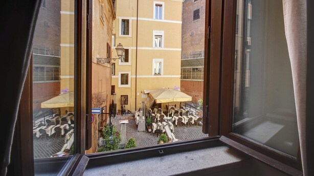 Vista dalla finestra di un edificio su un cortile. Si vedono tavoli e sedie all'aperto e un edificio con facciate giallo-marrone.