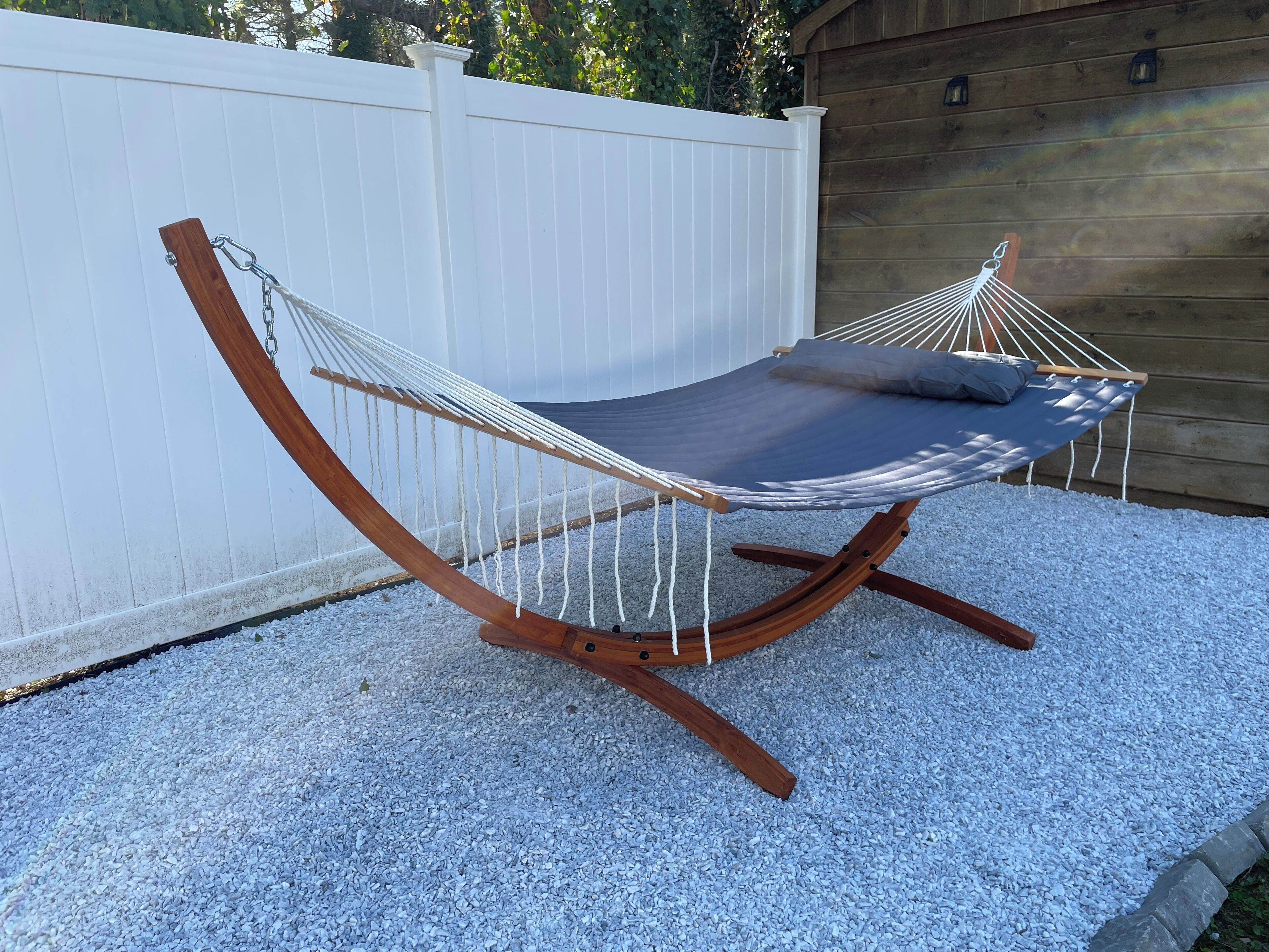 A hammock chair with a wooden frame and a grey cushion, set on a gravel surface with a white fence and wooden wall in the background.