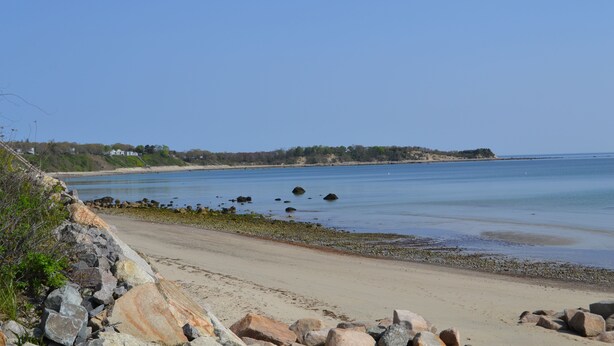 A beach with a rocky shoreline, clear blue water, and a distant view of houses.