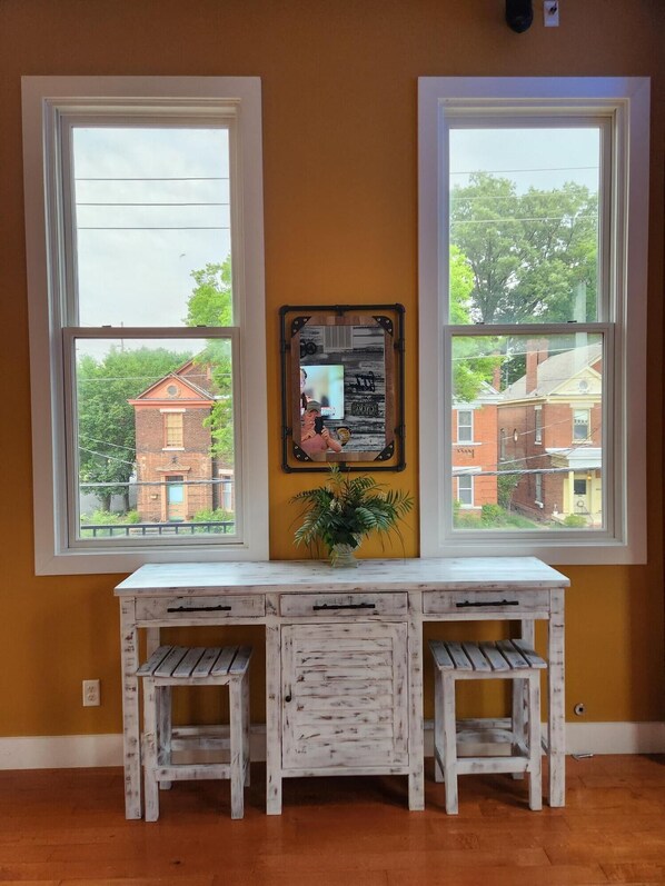 A room with a white desk and two stools, a framed picture on the wall, and a view of buildings outside through two windows.