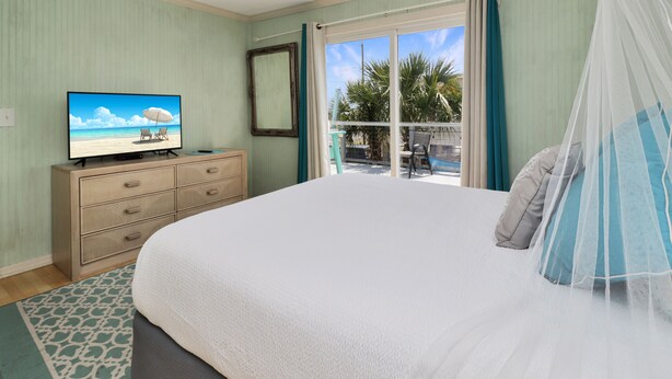 A bedroom with a bed, a television on a dresser, a window with curtains, and a view of palm trees outside. - Pet Friendly , Beach Views on the Quiet East End of SJB ~ Sea Star (St. Joe Beach)