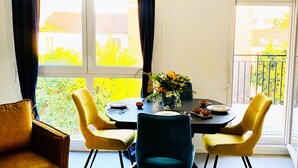 A dining area with a table set for four, surrounded by yellow chairs, a vase of flowers, and a view of a garden and buildings through large windows. - Luxury just 10 minutes from central Paris. Free Parking (Vitry-sur-Seine)