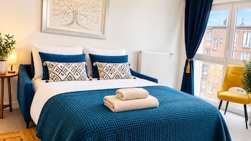 A bedroom with a bed, a window with curtains, a chair, a radiator, and a wall-mounted picture.