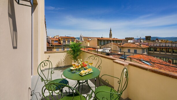 Un balcón con una mesa dispuesta para dos personas, con vistas a un paisaje urbano que incluye edificios históricos y un cielo azul despejado.