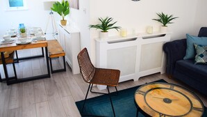 Un soggiorno moderno con un tavolo da pranzo in legno apparecchiato per quattro persone, un divano blu, un tavolino rotondo e due piante in vaso su un tavolo consolle bianco.
