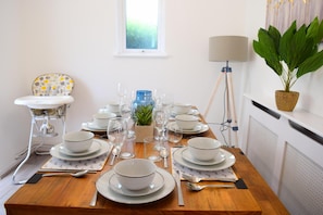 Uma mesa de jantar posta com pratos, tigelas e talheres brancos, uma pequena planta em vaso e um abajur.