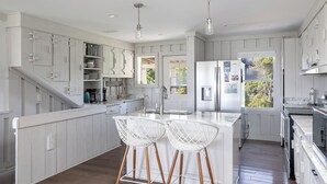 A modern kitchen with white cabinetry, a central island with bar stools, a refrigerator, and a window overlooking greenery. - Windy Dunes, Almost Oceanfront House in Emerald Isle, NC! (Emerald Isle)