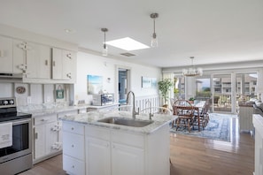 A modern kitchen with white cabinetry, a marble countertop, and a dining area with wooden chairs. - Windy Dunes, Almost Oceanfront House in Emerald Isle, NC! (Emerald Isle)