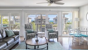 A spacious living room with a leather sofa, patterned armchairs, a round coffee table, and a ceiling fan. Large windows offer a view of the ocean and other buildings. - Windy Dunes, Almost Oceanfront House in Emerald Isle, NC! (Emerald Isle)
