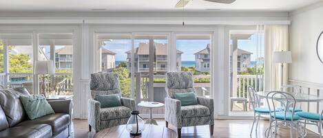 A spacious living room with a leather sofa, patterned armchairs, a round coffee table, and a ceiling fan. Large windows offer a view of the ocean and other buildings.