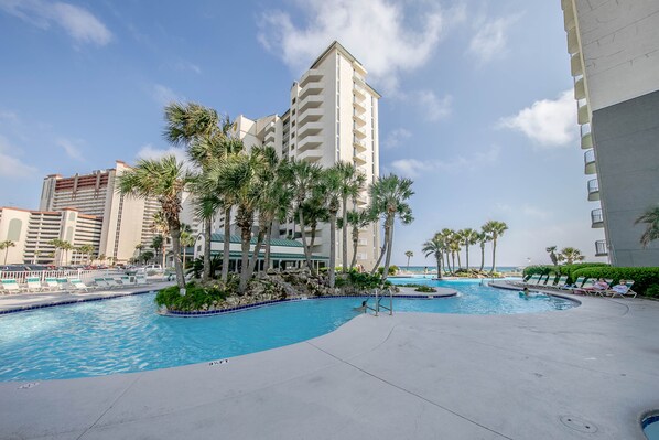 A resort pool area with palm trees, a clear sky, and a large building in the background.