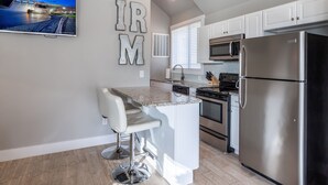 A modern kitchen with white cabinets, a stainless steel refrigerator, a microwave, and a built-in oven. There is a bar area with a white bar stool and a flat-screen TV mounted on the wall displaying a scenic view.