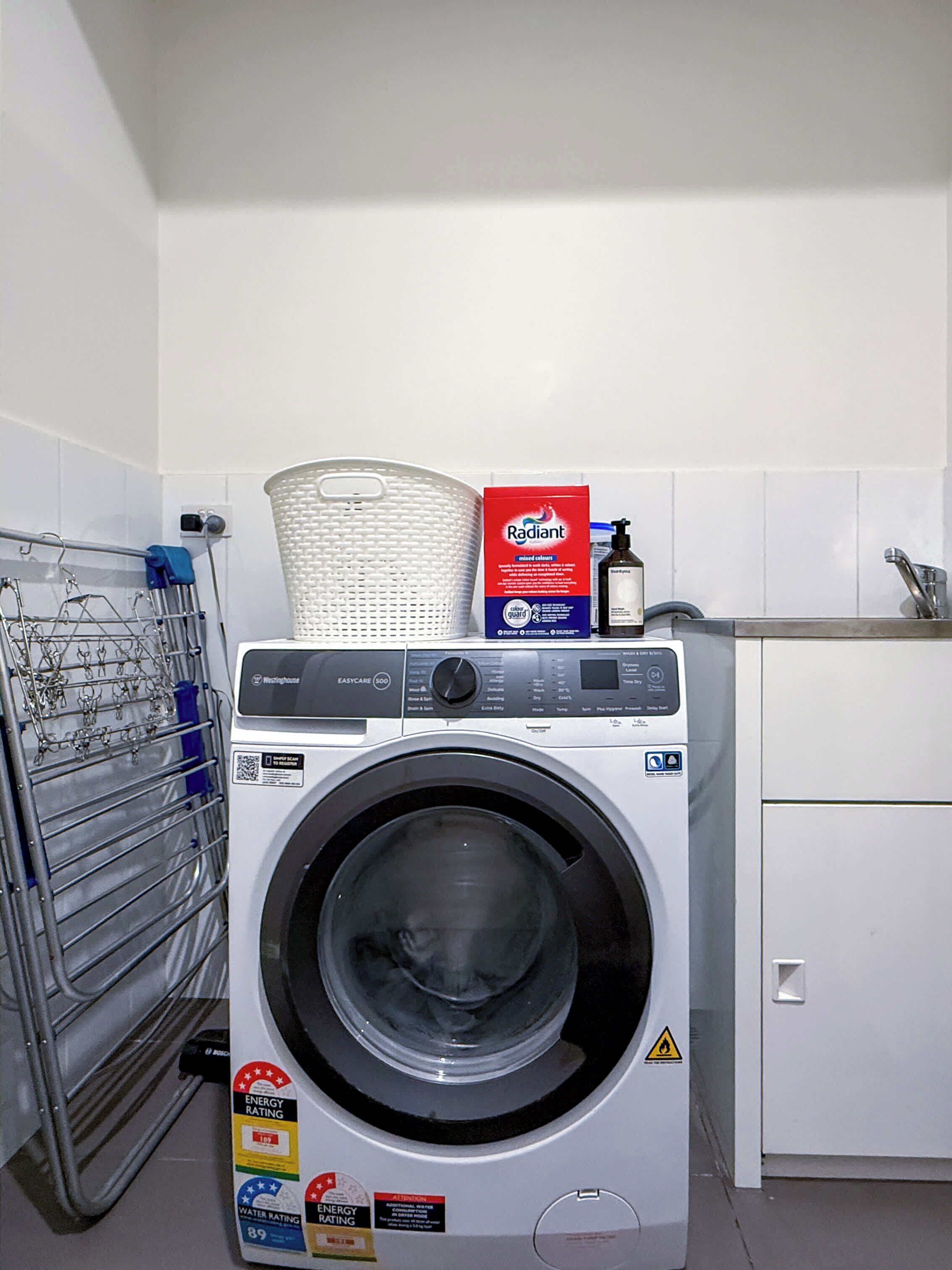 A front-loading washing machine with a basket on top, a box of detergent, and a rack of hanging clothes.