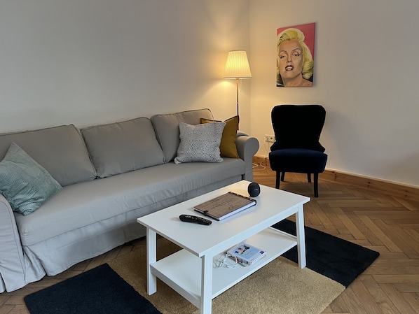 Ein Wohnzimmer mit grauem Sofa, weißem Couchtisch, schwarzem Sessel, einer Lampe und einem Bild an der Wand.