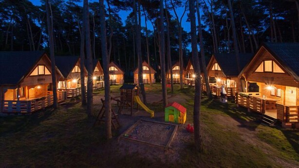 Une rangée de cabanes en bois dotées de grandes fenêtres, entourées de grands arbres, et une aire de jeux au centre.