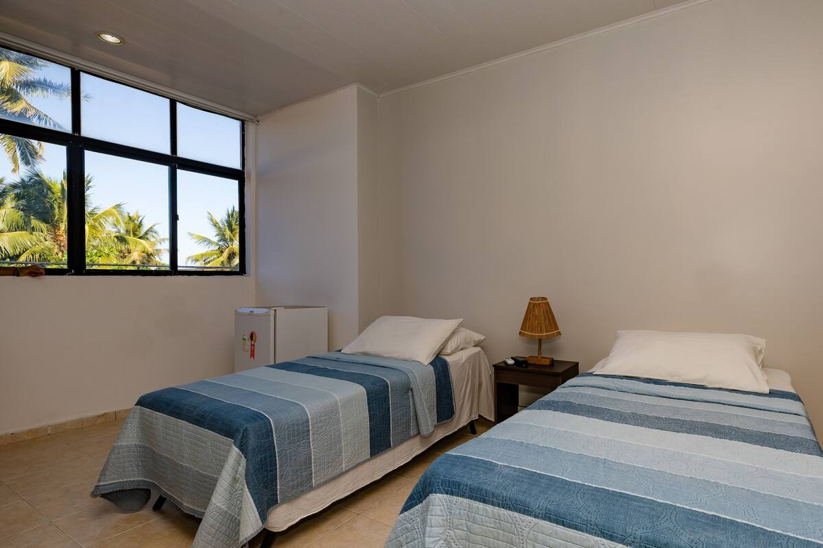 A room with two beds, a window with a view of palm trees, a small table with a lamp, and a refrigerator.