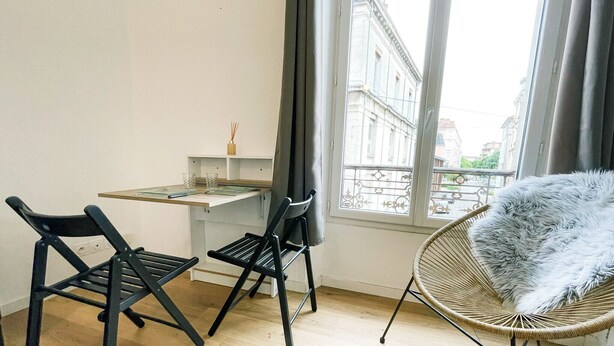 Una habitación moderna con escritorio, dos sillas negras, una silla de mimbre con un cojín mullido y vistas a edificios a través de amplios ventanales.