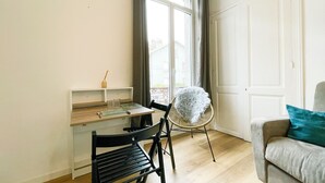 Una habitación con un escritorio de madera, una silla negra, una silla de mimbre, un sofá gris con un cojín azul turquesa y una ventana con cortinas.