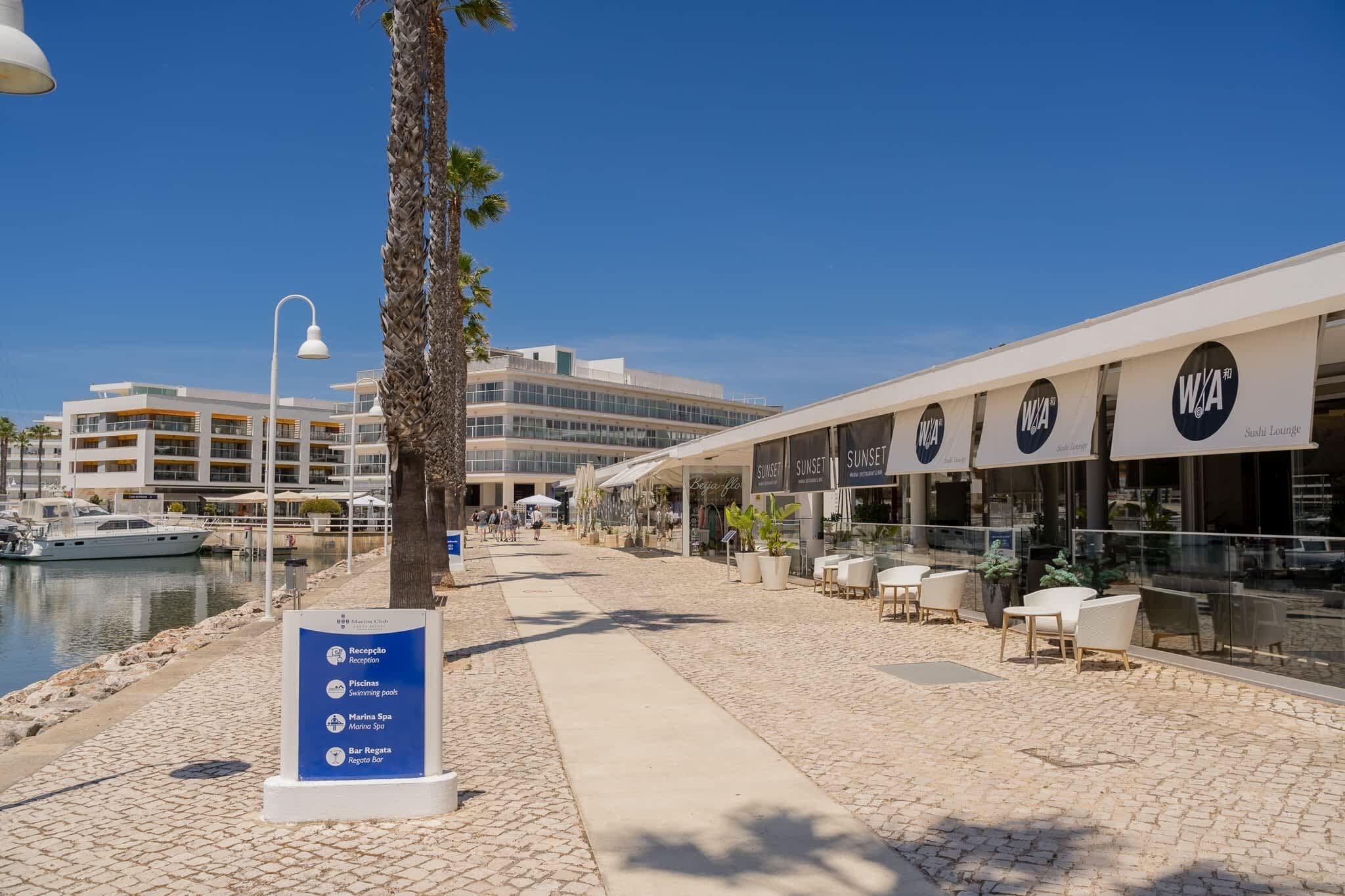 A waterfront promenade with a marina, a modern building with balconies, and a row of outdoor seating areas with umbrellas.