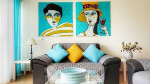 A living room with a sofa, two cushions, a glass coffee table, a lamp, and two paintings on the wall.