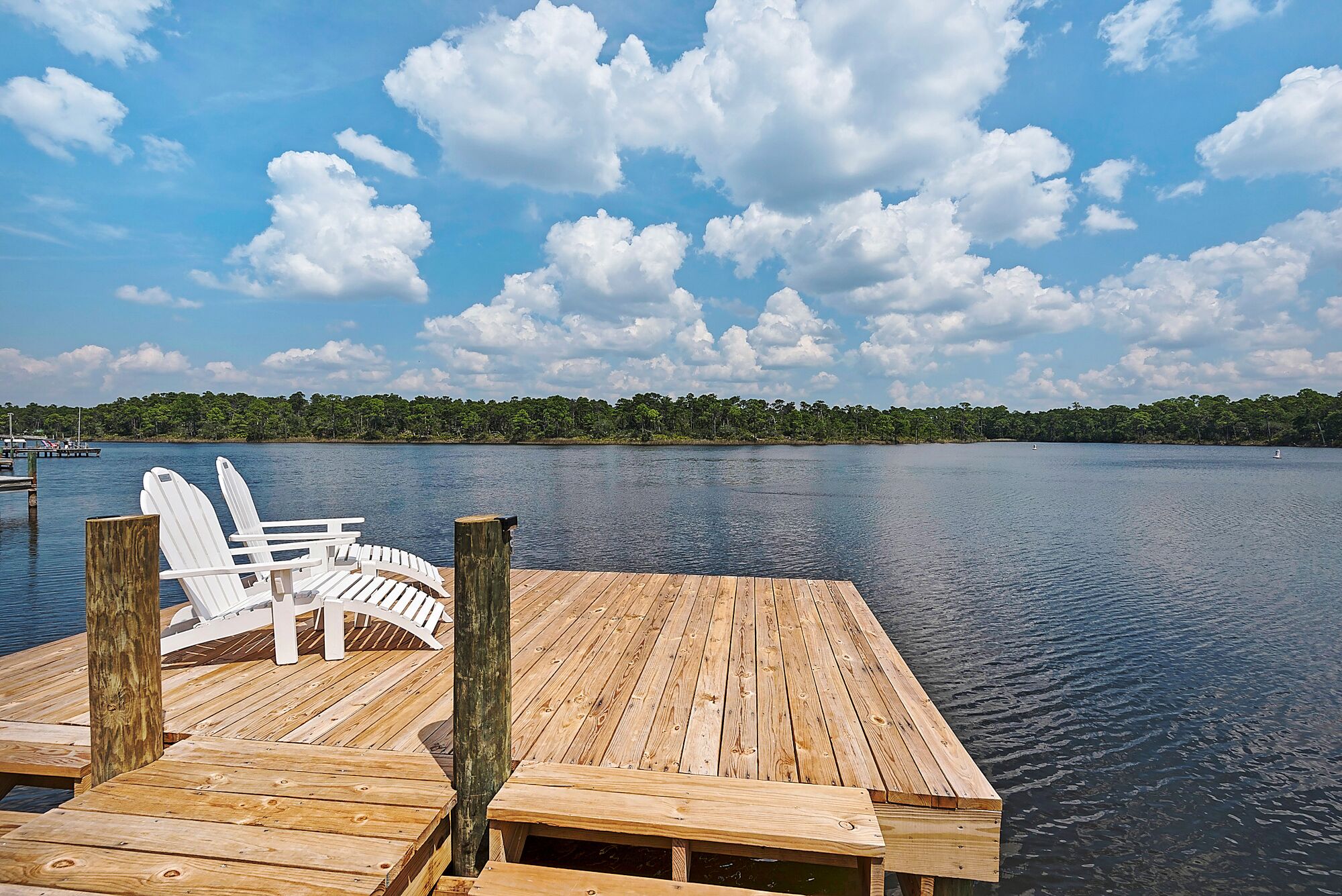 A wooden dock with white Adirondack chairs and a view of a calm lake with a forested shoreline.