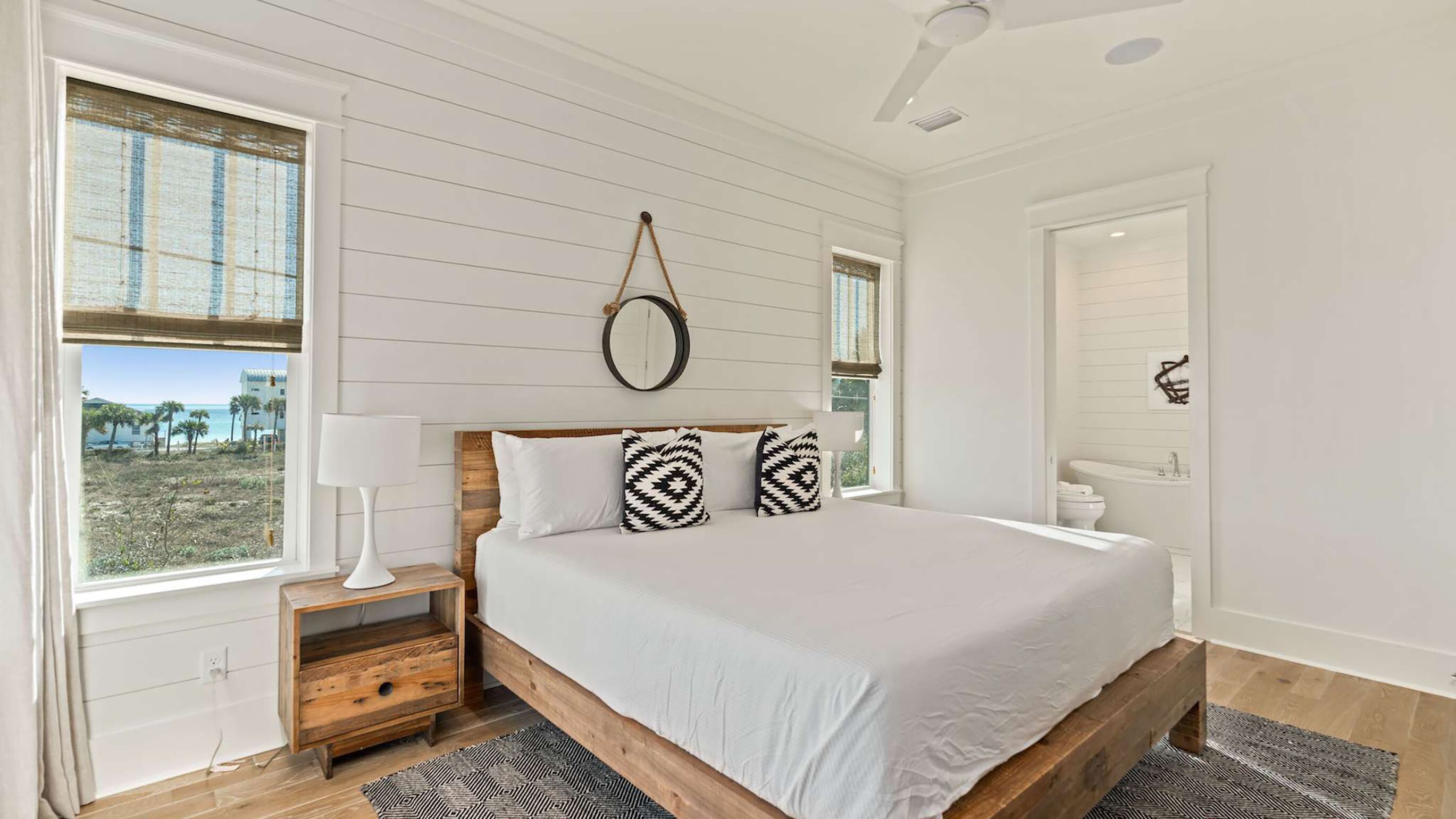 Een slaapkamer met een groot bed, houten nachtkastjes, een spiegel en uitzicht op het strand.