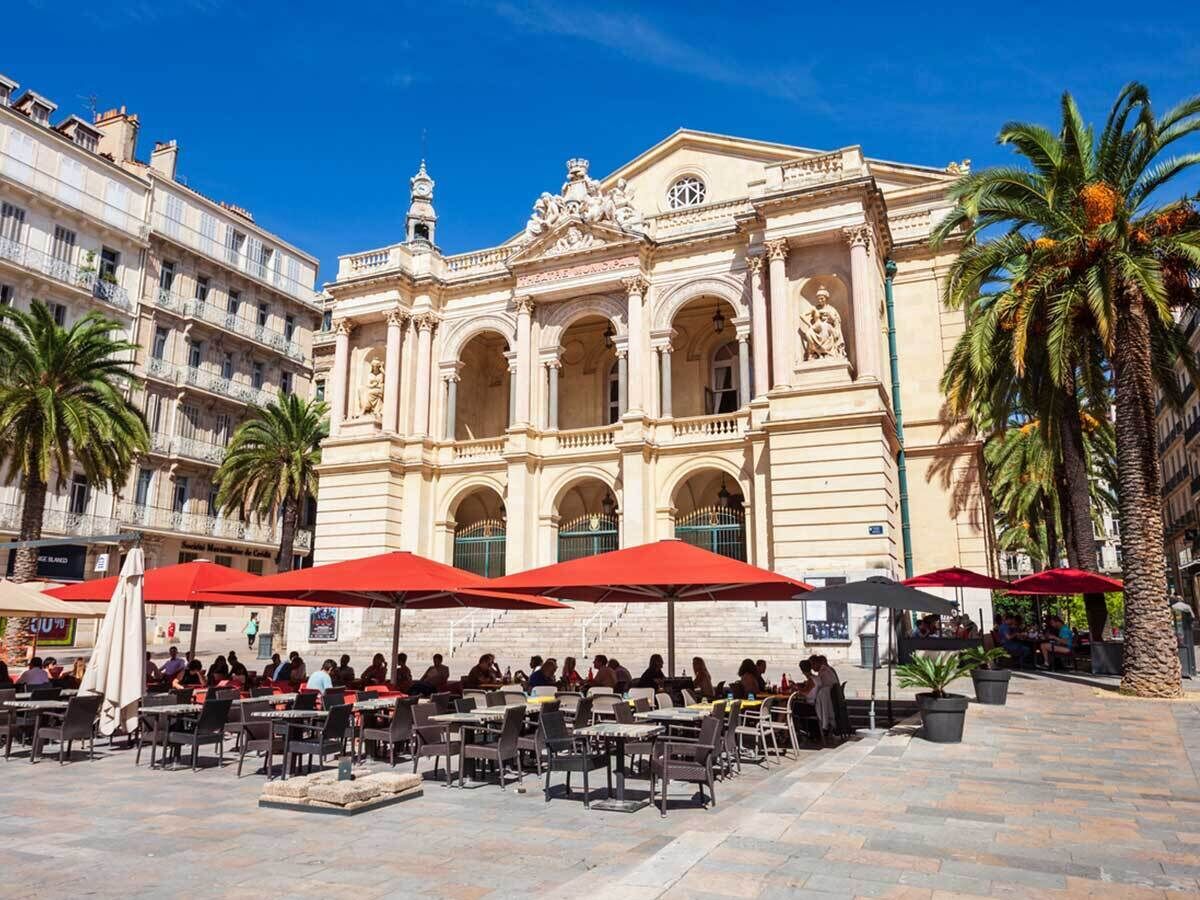 An outdoor café with tables and red umbrellas in front of a historic building with ornate architecture.