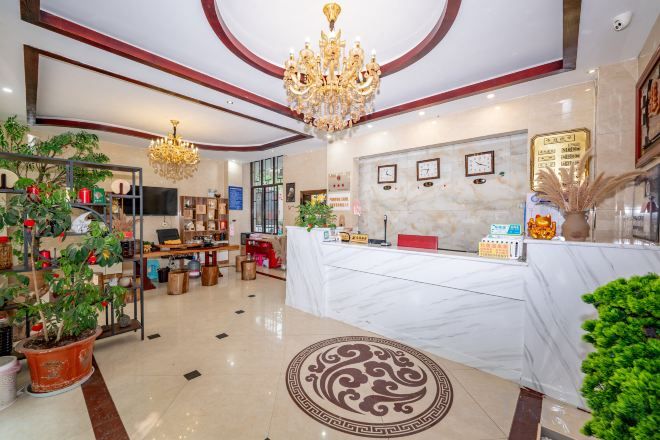 A hotel lobby with a reception desk, a chandelier, potted plants, and a marble floor.