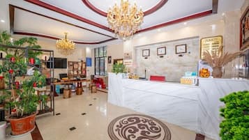 A hotel lobby with a reception desk, a chandelier, potted plants, and a marble floor.