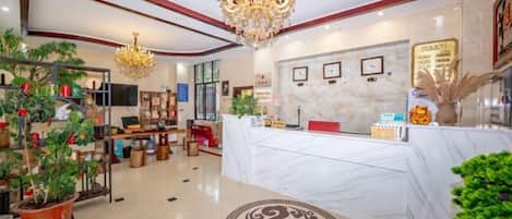 A hotel lobby with a reception desk, a chandelier, potted plants, and a marble floor.