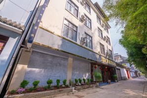 A multi-story building with a traditional architectural style, featuring a large entrance with a covered awning and a row of small plants along the walkway. - Qianshuiwan Hotel (Tengchong)