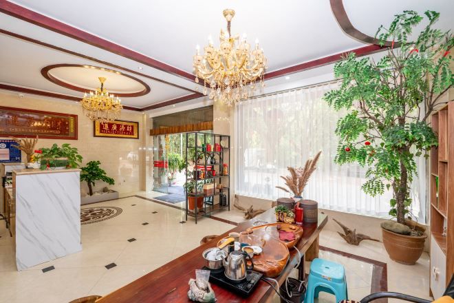 A hotel lobby with a reception desk, a chandelier, potted plants, and a large window with curtains.